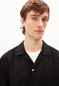 Zwart shirt met korte mouwen, met een notch-kraag, een enkele borstzak en een witte knop. Gemaakt van textuurstof, gelaagd over een wit t-shirt.