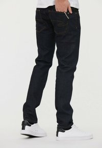 Vaqueros de mezclilla oscura con corte slim, costuras amarillas en contraste y bolsillos traseros con un diseño curvado. Combinados con zapatillas blancas.