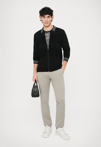 Cardigã preto com fecho éclair e gola às riscas, usado sobre uma camisa às riscas, combinado com calças bege e ténis brancos. Modelo segurando uma mala preta.