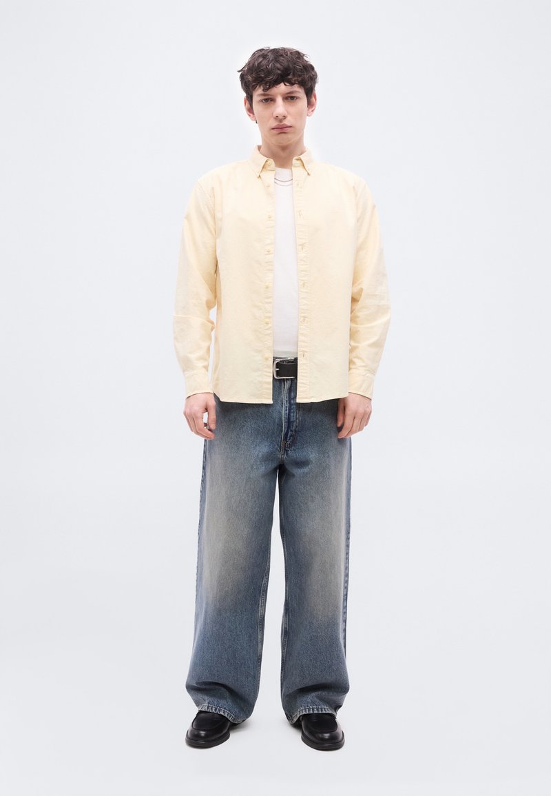 Jeune homme debout portant une chemise jaune pâle à boutons sur un tee-shirt blanc, un jean bleu délavé et ample, une ceinture noire et des chaussures noires sur fond blanc.