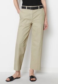 Beige katoenen broeken met een rechte pijp, voorzien van zijzakken en een zwarte riem. Gepaard met zwarte slip-on sandalen op een neutrale achtergrond.