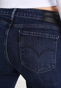 Mörkblå denimjeans med en bakficka som har ett subtilt V-mönster och ett märkesmärke i svart läder vid midjan.
