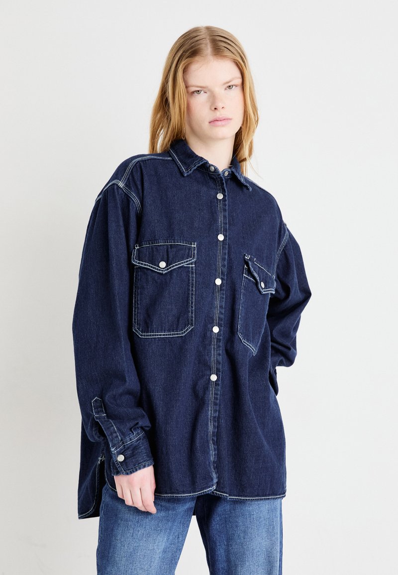 Mørkeblå denimskjorte med krave, knaplukning, to brystlommer og hvide syninger. Løs pasform med lange ærmer.
