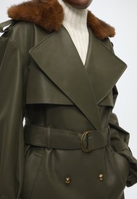 Trench-coat en cuir vert olive avec un col en fourrure marron, de larges revers, une taille ceinturée et des boutons d'accent dorés à l'avant.