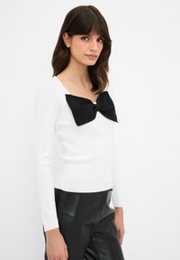 Weißes Langarmshirt mit quadratischem Ausschnitt, versehen mit einer großen schwarzen Stoffschleife an der Vorderseite. Glatte Textur, figurbetonter Schnitt.