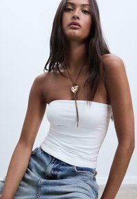 Vit axelbandslös topp, jeansshorts, brunt halsband med hjärtberlock. Långt mörkt hår, naturlig makeup, sittande pose, neutral bakgrund.