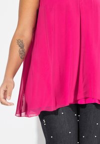 Top sin mangas de un brillante color rosa, con un diseño fluido y aberturas laterales, combinado con unos pantalones oscuros decorados con cuentas blancas.