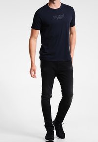 Muž oblečený v námořnické modré košili s krátkým rukávem s textem, černých slim-fit džínech a černých teniskách, stojící proti bílému pozadí.