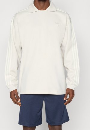 Muž oblečený v krémové mikině s límečkem a dlouhými rukávy, s jemným logem a pruhy na rukávech, kombinovanou s tmavě modrými šortkami.