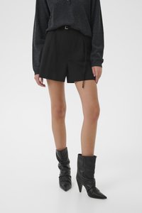 Persona que lleva pantalones cortos negros de tiro alto, una camiseta de manga larga gris oscuro y botines negros de tacón afilado sobre un fondo liso.