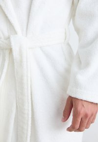 Accappatoio in spugna bianca con una superficie testurizzata, dotato di cintura in vita e maniche lunghe, con una mano che afferra l'accappatoio.