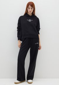 Zwarte hoodie met bijpassende joggingbroek. De hoodie heeft een witte grafische tekst; de broek heeft een trekkoord. Gladde stof en een ontspannen pasvorm.