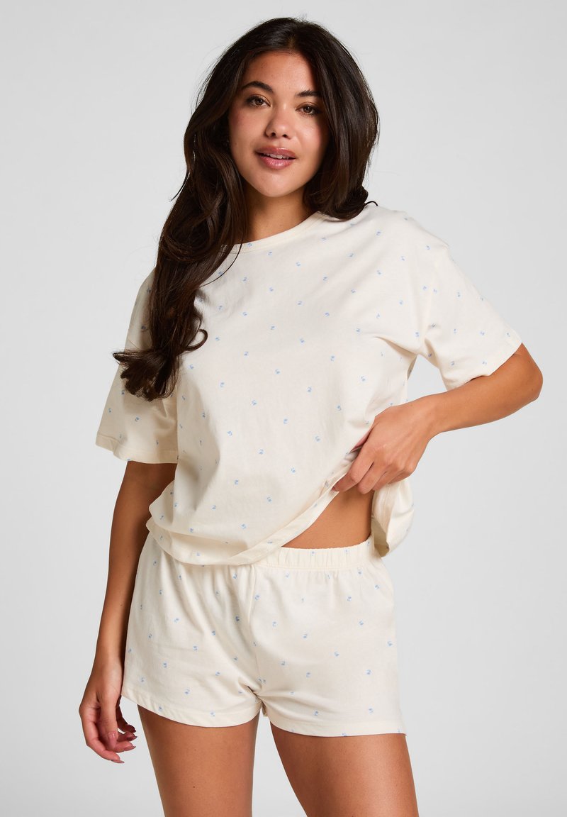 Young woman with long dark hair wearing a cream short-sleeve top and matching shorts with small blue dots, lifting her top slightly.