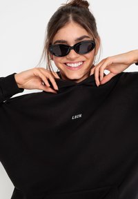 Felpa nera con cappuccio e tasca frontale, con il logo "LSCN" in bianco. Il modello indossa occhiali da sole neri evela una texture del tessuto liscia e una vestibilità rilassata.