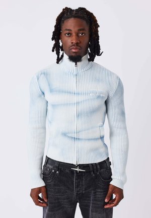Homme aux cheveux tressés portant un pull zippé côtelé bleu clair et blanc et un jean noir avec une ceinture noire sur un fond blanc uni.
