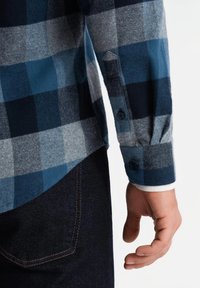 Camisa de franela a cuadros azul y gris con puños abotonados, material interior blanco en contraste y línea de dobladillo estructurada. Se ven jeans oscuros.