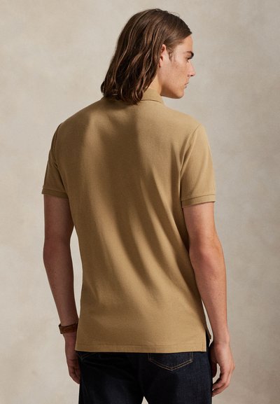 Mladi moški z rjavimi ramenskimi lasmi, oblečen v bež polo majico in temne kavbojke, viden od zadaj na nevtralnem ozadju.