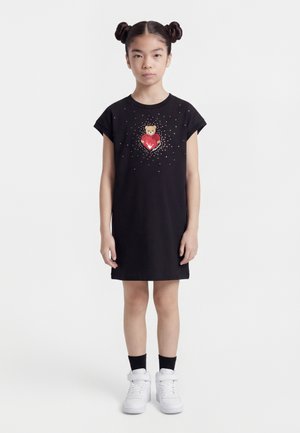 Dievča s dvojnásobnými drdolmi, oblečené v čiernych šatách s potlačou medvedíka držiaceho červené srdce obklopené bielymi bodkami, v kombinácii s bielymi teniskami a čiernymi ponožkami.