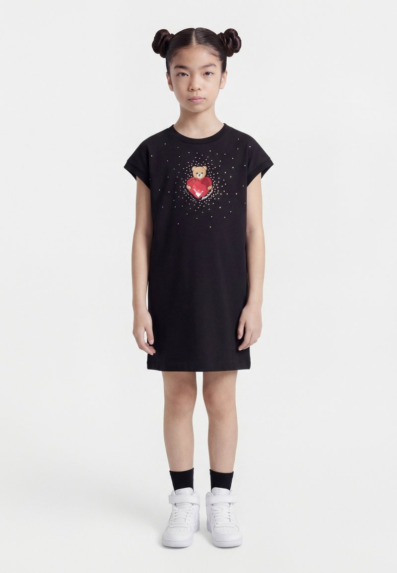 Κορίτσι με διπλούς κότσους που φοράει μαύρο φόρεμα με σχέδιο ένα αρκουδάκι που κρατάει μια κόκκινη καρδιά περιβαλλόμενη από λευκές τελείες, συνδυασμένο με λευκά αθλητικά παπούτσια και μαύρες κάλτσες.