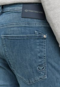 Blauwe denim jeans met een standaard achterzak, zilveren knoopdetail en een zwart leren label met "TOM TAILOR" gedrukt.