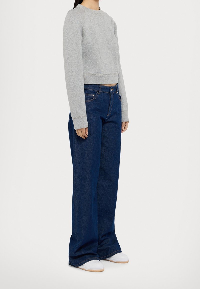 Personne portant un sweat court gris à manches longues, un jean large bleu foncé et des baskets blanches, debout sur un fond blanc.