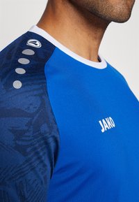 Maglietta sportiva blu a maniche corte, colletto bianco, pannelli laterali di un blu scuro strutturato e logo "JAKO" bianco al centro davanti.