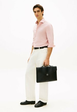 Ung mand iført en lyserød skjorte, hvide bukser, sort bælte og sko, der holder en sort lædermappe mod en ensartet hvid baggrund.