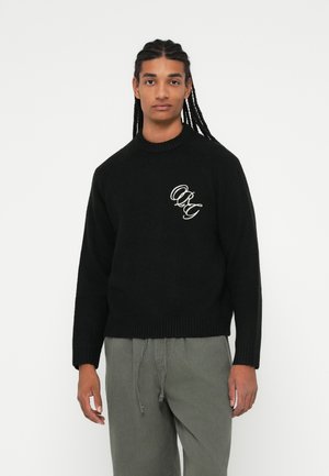 Black knitted sweater with a ribbed neckline and cuffs, featuring a white embroidered logo on the front. Worn with gray pants.