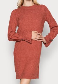 Kopparfärgad tröjkklänning med hög hals, långa ärmar, texturerad tyg och knappar på ärmarnas manschetter. Enkel, rak siluett.