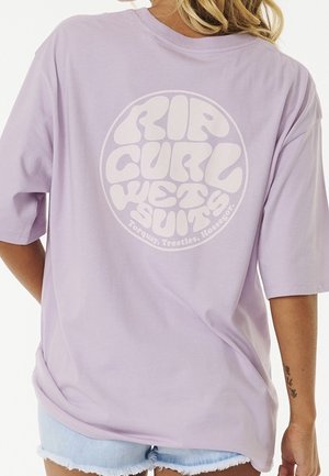 Persoon draagt een lavendelkleurig Rip Curl wetsuit t-shirt en lichtblauwe denim shorts, met een tatoeage op de linker onderarm zichtbaar.