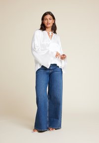 Hvit, flytende bluse med vide ermer og knyting i halsen, sammen med blå vide jeans. Glatt tekstur, minimalistisk design.