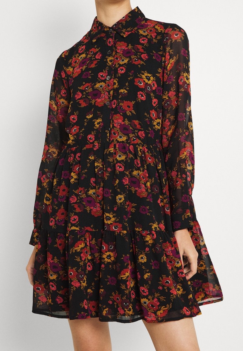 Robe noire à manches longues avec motif floral rouge, violet et jaune, boutonnée à l'avant et jupe à volants portée par une personne debout.