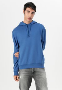 Modra kapuca s vrvico in kenguru žepom, z ribanim zaključkom na rokavih in spodnjem robu. Nosijo se z zbledelimi sivimi jeans hlačami.