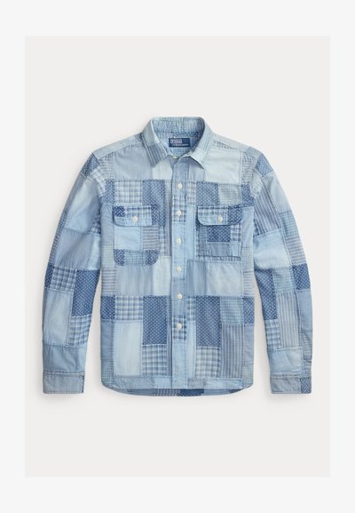 Chemise patchwork en denim bleu clair à manches longues, avec fermeture à boutons sur le devant et deux poches poitrine à boutons.