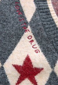 Grå och krämfärgad stickad tyg med en röd stjärna och texten "LOVE IS THE DRUG" broderad i rött. Subtil textur synlig.