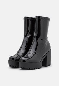 Bottines en cuir verni noir avec talons hauts épais, fermeture éclair à l'avant et semelles à plateforme rainurées sur fond blanc.