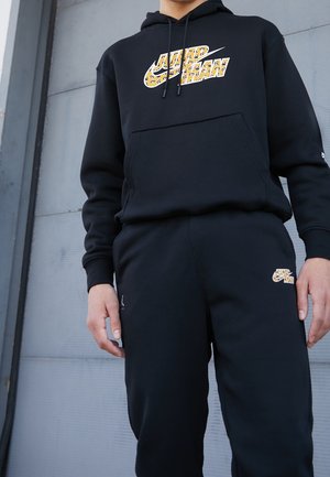 Sudadera con capucha y pantalones deportivos negros con un logo amarillo con patrón en el pecho y un logo más pequeño en el muslo izquierdo.