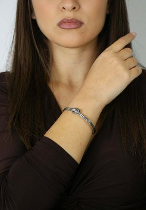 Srebrna bransoletka o splecionej konstrukcji z centralnym detalem węzła, noszona na nadgarstku o jasnym odcieniu skóry. Tło jest neutralne.