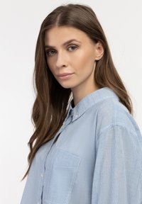 Camicia a righe azzurro chiaro con colletto, taschino sul petto singolo e chiusura anteriore con bottoni. Presenta una texture morbida e una vestibilità comoda.