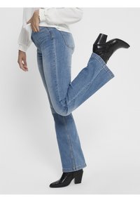 Nainen, joka käyttää vaaleansinisiä farkkuja ja mustia korokepohjaisia nilkkasaappaita, nostaa toista jalkaansa polvesta koukussa valkoista taustaa vasten.