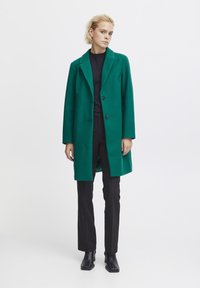 Green wool-blend coat with a notched collar, two black buttons, and a relaxed fit, paired with black trousers and ankle boots.