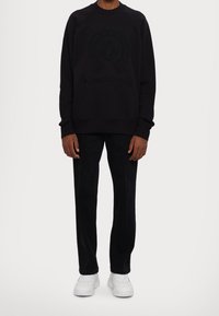 Schwarzer Pullover mit geprägten Logodetails, kombiniert mit schwarzen Hosen und weißen Sneakers. Glatte Textur, lockere Passform und Rundhalsausschnitt.