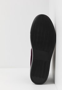 Podeszwa czarnego kauczukowego buta z falistym wzorem bieżnika, ustawiona pionowo wzdłuż białej ściany.