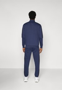 Fato de treino azul-marinho com um top de gola com fecho e calças de fato correspondentes. Apresenta realces pretos ao longo das laterais e punhos elásticos. Ténis brancos visíveis.