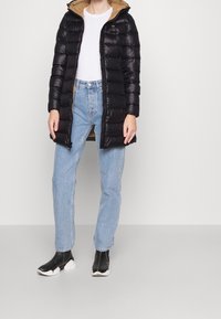 Sort pufferjakke med en kontrasterende brun inderside, lang længde, lynlåslukning og vandret quiltet mønster, båret med lyseblå jeans og sorte støvler.