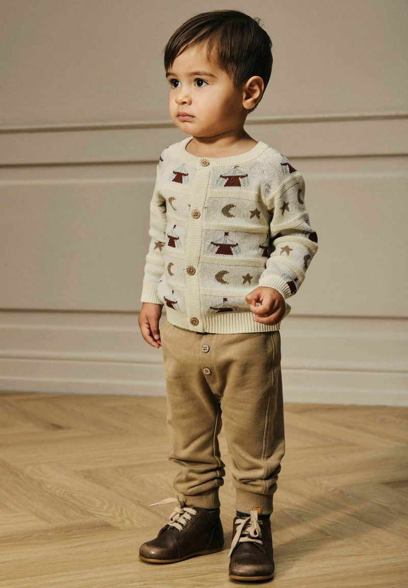 Beige stickad cardigan med röda och bruna mönster, kombinerad med ljusbruna byxor och mörkbruna snörskor.
