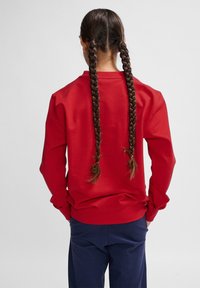 Felpa rossa a maniche lunghe con vestibilità rilassata, scollo rotondo e polsini, abbinata a pantaloni blu navy. Capelli acconciati in due trecce.