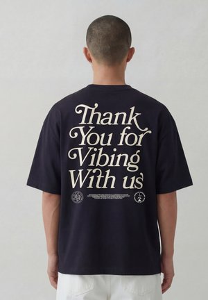 Persoon met kort haar draagt een zwart T-shirt met grote witte tekst op de achterkant waarop staat: "Thank You for Vibing With us".