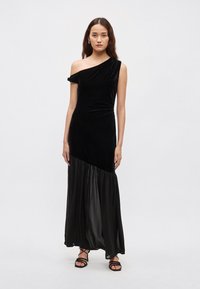 Czarna aksamitna sukienka z asymetrycznym, jedno-ramiennym krojem, opadająca od dopasowanego gorsetu do plisowanej, szyfonowej spódnicy. Czarne sandały na paski.