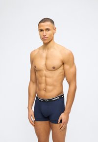 Μυώδης άνδρας με κοντά μαλλιά, φοράει μπλε σκούρα boxer της Nike, στέκεται μπροστά σε φωτεινό γκρι φόντο, αντιμετωπίζοντας προς τα εμπρός.